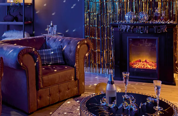 Wohnzimmer mit goldenem Lamettavorhang, einem Kamin mit einer Happy-New-Year-Girlande, 1 brauner Ledersessel und Couchtisch mit goldfarbenem Gestell.