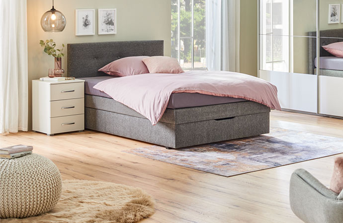 Graues Boxspringbett, rosa Bettwäsche, weißer Nachttisch, weißer Schrank mit Spiegelfront in einem Schlafzimmer