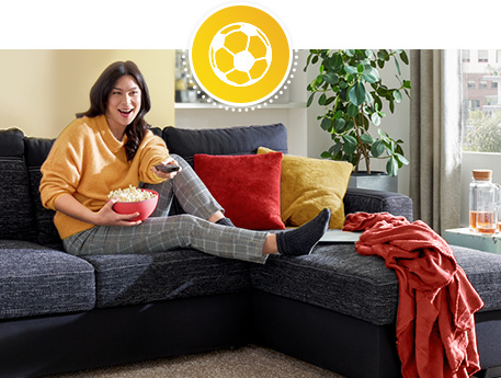 Frau auf dunkelgrauem Sofa sitzend, rote, gelbe Kissen, rote Decke, Fußball im Regal, weitere Deko.