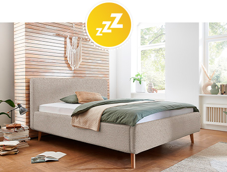 Schlafzimmer mit beigem Bett mit grüner Bettwäsche und beigem Plaid vor Holzwand und weißer Wand mit großen Fenstern