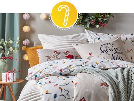 Bett mit diversen weihnachtlichen Bettwöschen mit Lichterkette und Hunden als Druck sowie Print "A house is not a home without a dog", hellgrüne Decke, Kissen mit Dackel, gelbes Kissen, weißes Kissen, Beistelltisch aus Holz mit Vase samt Zweigen und Geschenk.