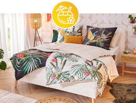 Schlafzimmer mit weißem Bett, schwarzer und weißer Wendebettwäsche mit einer Uniseite und einer Seite mit floralem Muster, Papageien- und Palmen-Druck, Sisal-Teppich, Beistelltisch aus Rattan, Rattan-Leuchte