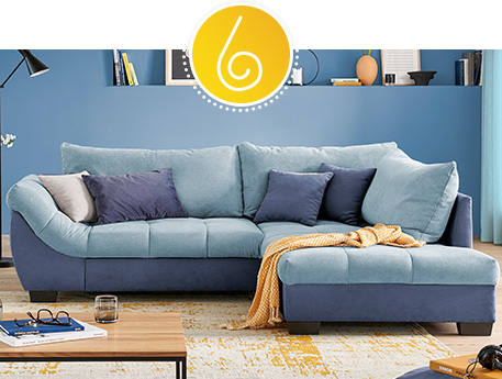 Ecksofa in verschiedenen Blautönen mit passenden Kissen, gelb-weißer Fransendecke, beige-gelber Teppich, blaue Wand mit Deko