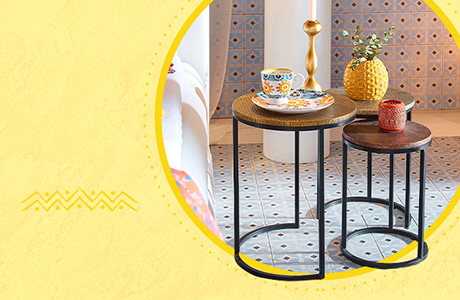 Raum im Ethno-Stil mit 3 Beistelltischen unterschiedlicher Größe mit Tischplatten in Gold, Silber und Bronze, Geschirr, Kerzenstönder und Vasen im Ethno-Stil, blau gemusterte Fliesenwand und -boden